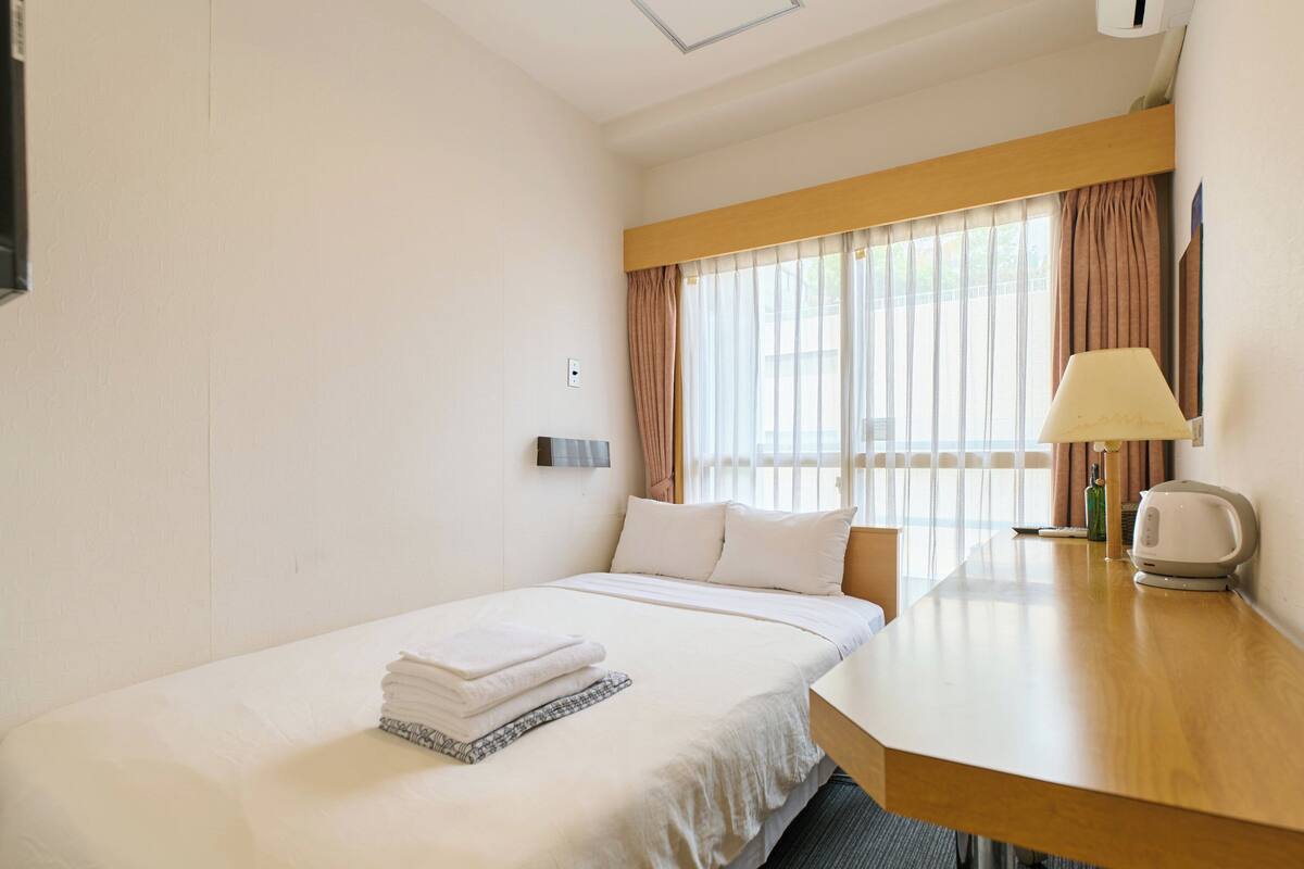 A hotel room with a single bed, a bedside table with a lamp, a window with curtains, and a wall-mounted light switch.