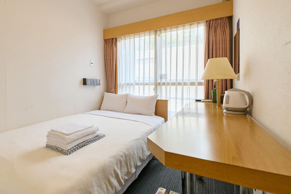 Una habitación de hotel con cama, escritorio, lámpara y ventana con cortinas.