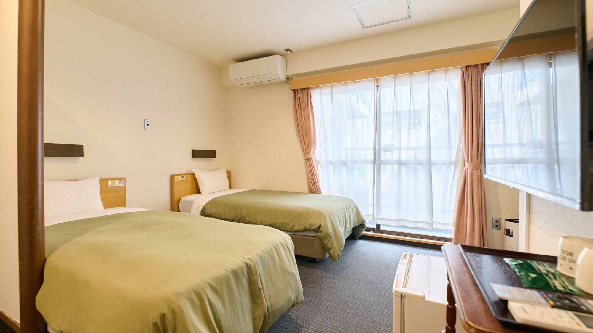 Hotelkamer met twee bedden, een bureau met een computer, een raam met gordijnen en een airconditioner.