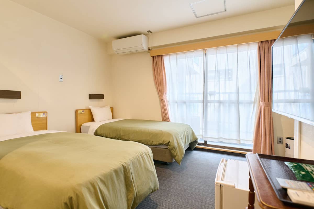 Habitación de hotel con dos camas, un escritorio con computadora, una ventana con cortinas y un equipo de aire acondicionado.