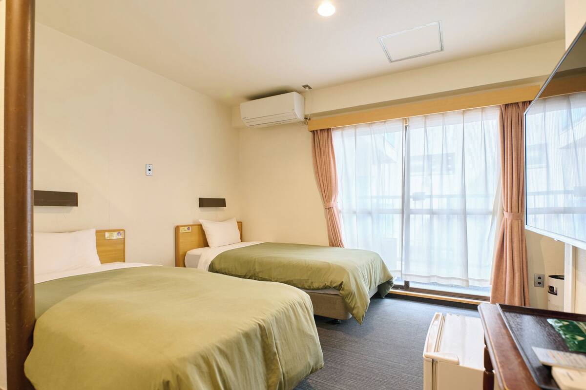 A hotel room with two single beds, a desk, a chair, an air conditioning unit, and a window with curtains.