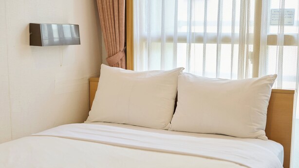 A neatly made bed with white linens and two pillows, a wall-mounted light fixture, and a window with sheer curtains. - Tabist Hotel Tetora Ikebukuro (Tokyo)