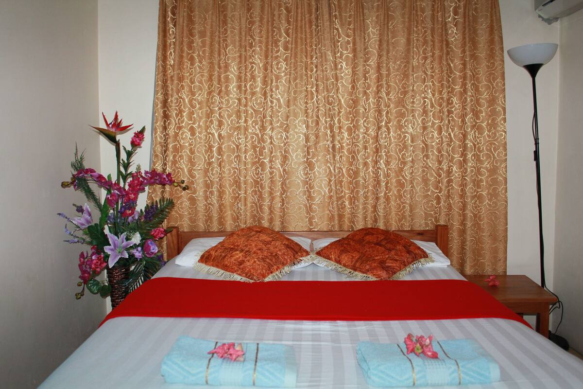 A neatly made bed with a red bedspread, two pillows, and a floral arrangement beside it.