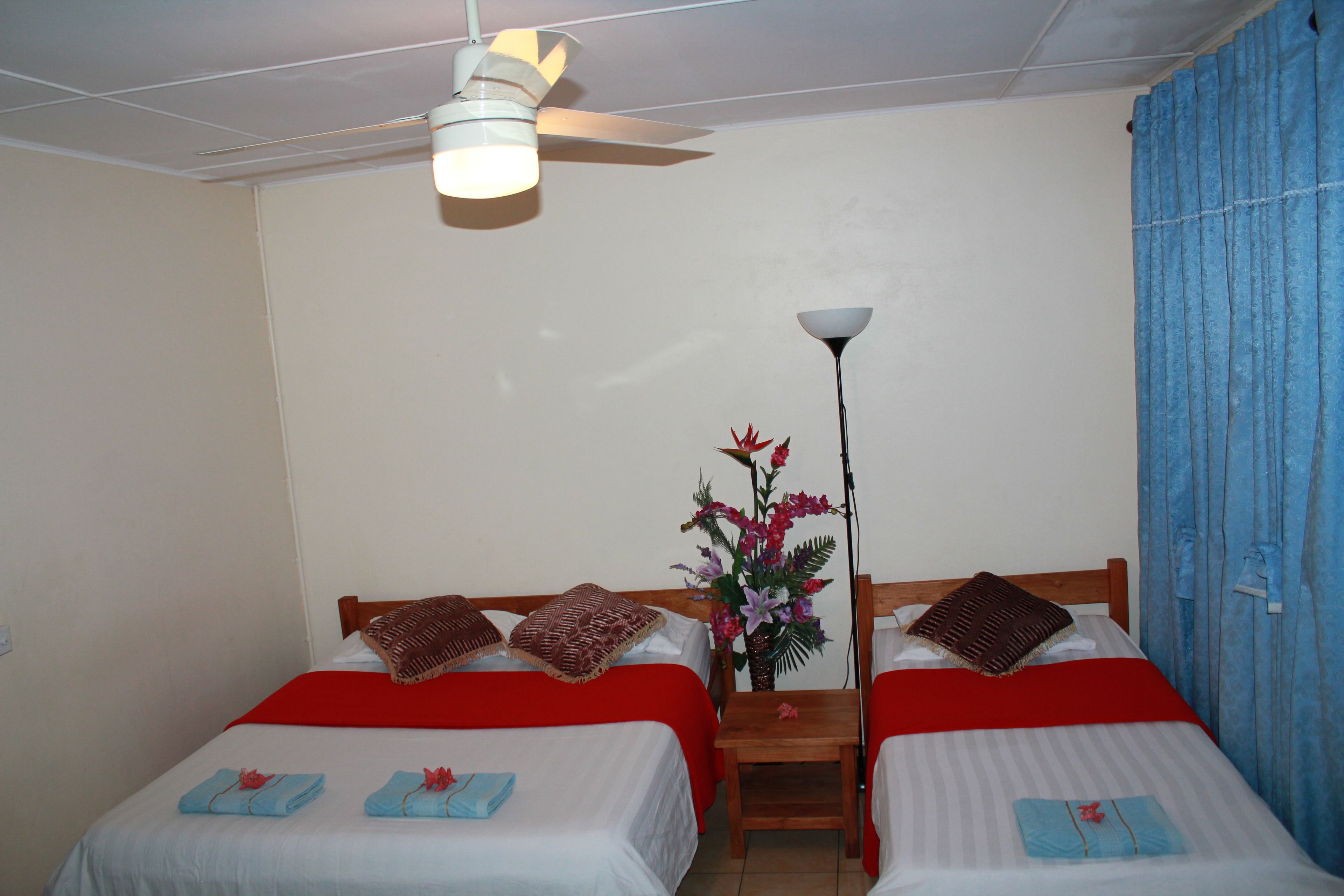 A room with two beds, each with a red blanket and pillows, a ceiling fan, a nightstand with a lamp, and a vase with flowers.