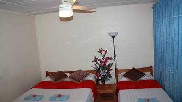 A room with two beds, each with a red blanket and pillows, a ceiling fan, a nightstand with a lamp, and a vase with flowers.