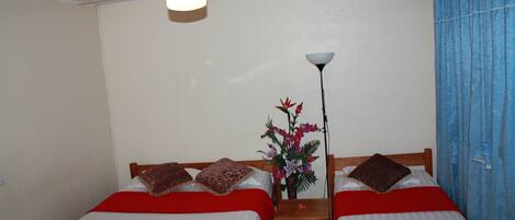 A room with two beds, each with a red blanket and pillows, a ceiling fan, a nightstand with a lamp, and a vase with flowers.