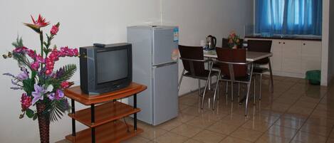 A small kitchenette with a refrigerator, a television on a wooden stand, a dining table with chairs, and a window with blue curtains.