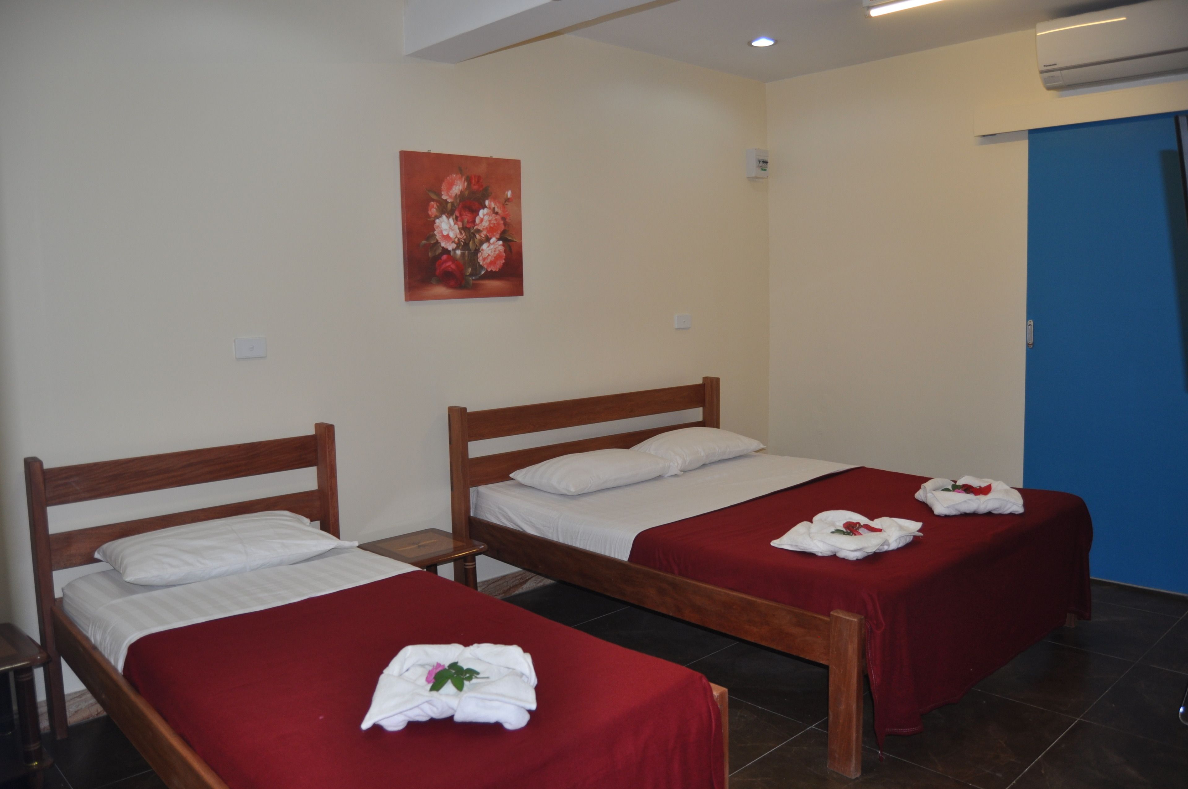 Habitación de hotel con dos camas individuales, cada una con una colcha roja y almohadas blancas. En la pared, hay un cuadro de flores, y un equipo de aire acondicionado también montado en la pared.