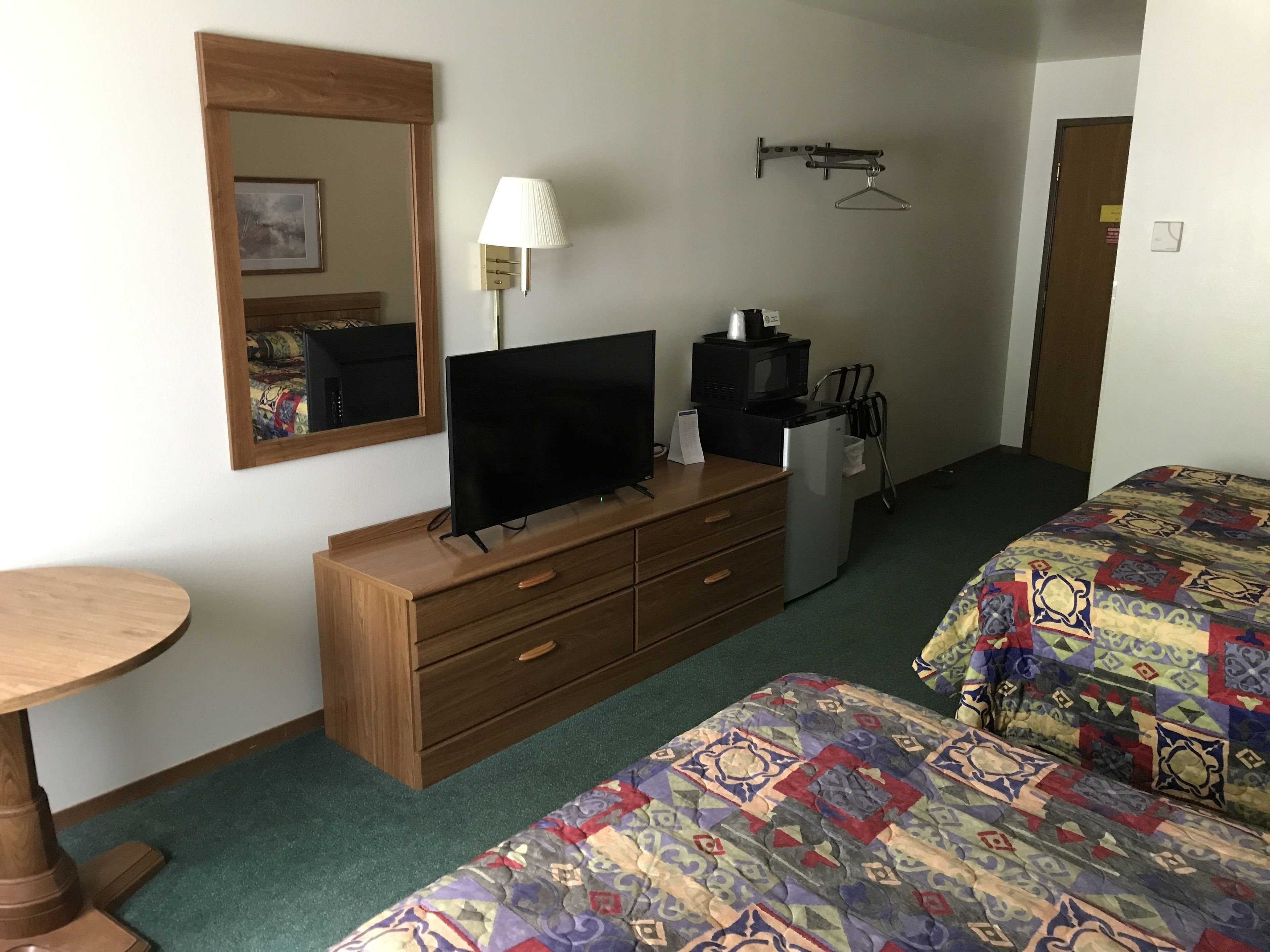 Ein Hotelzimmer mit einem Holzschrank, einem Fernseher, einem kleinen Kühlschrank, einer Mikrowelle, einem Kleiderständer und einem Bett mit einem gemusterten Überwurf, sowie einem runden Holztisch.