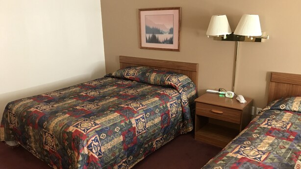 A hotel room with two single beds, a nightstand with a lamp, a framed picture on the wall, and a small table with a phone and a clock. - Badger Motel (Chippewa Falls)