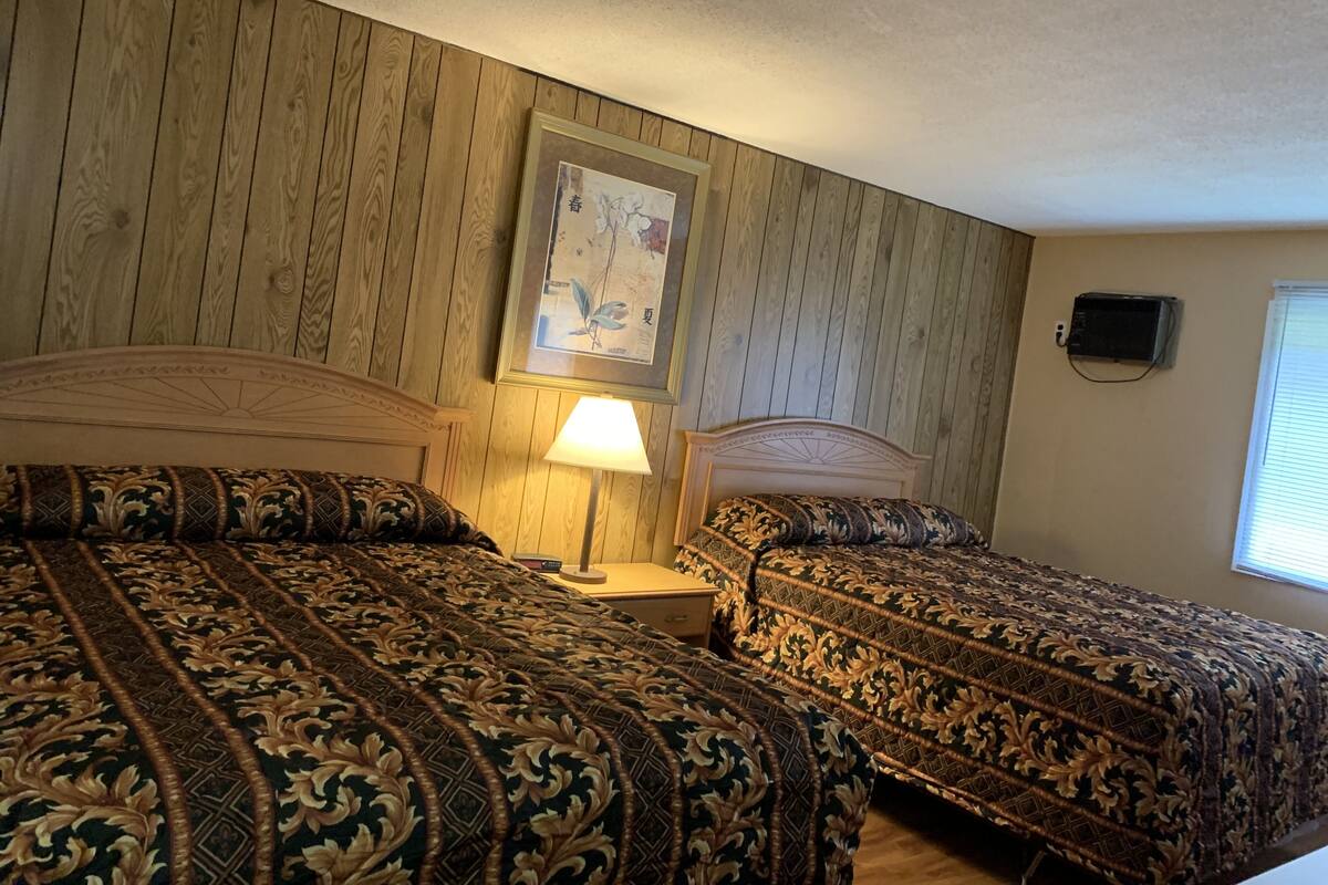 A hotel room with two beds, a nightstand, a lamp, a framed picture on the wall, and a window with blinds.