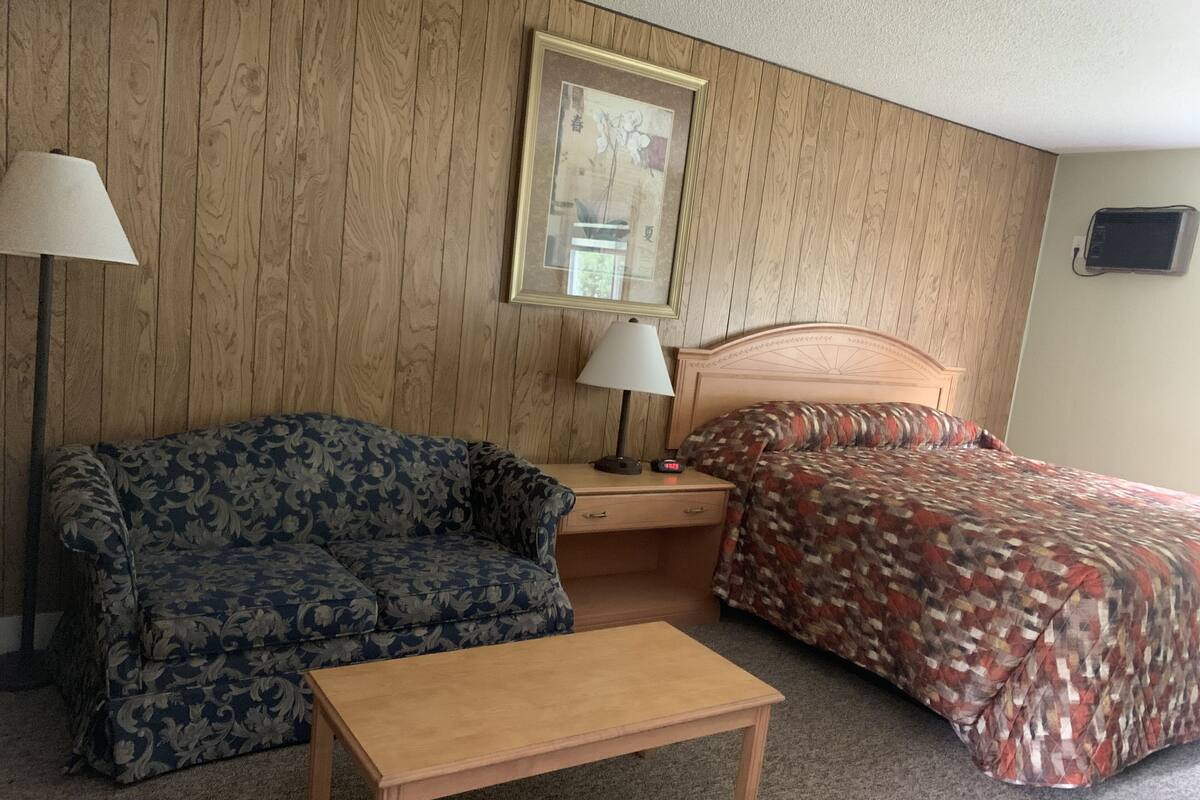 A room with a wooden panel wall, a sofa, a bed with a patterned bedspread, a bedside table with a lamp, and a small wooden table.