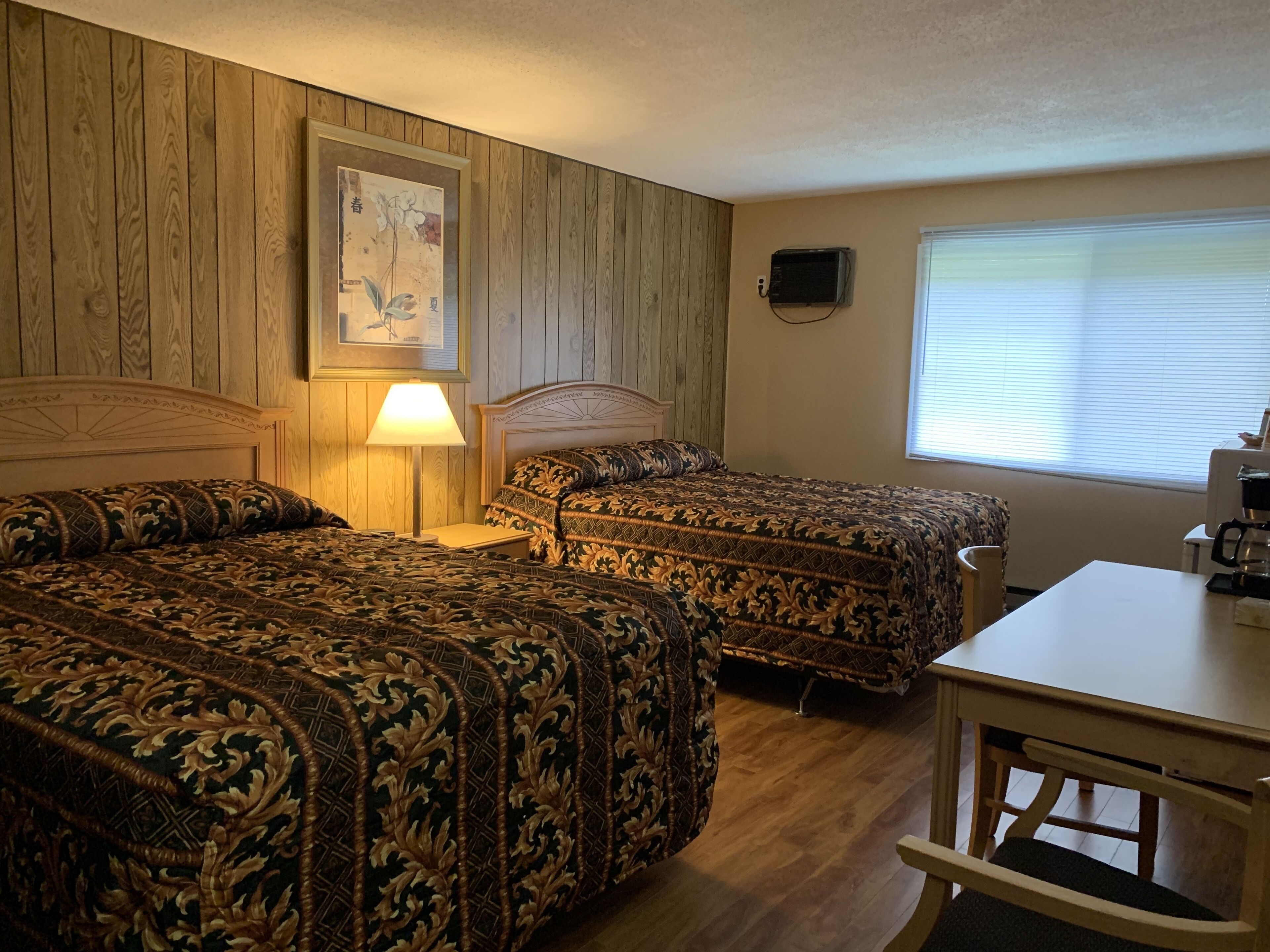Una habitación con dos camas, una mesa, una lámpara, un cuadro en la pared y un ventanal con persianas.
