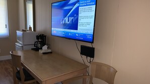 A room with a wooden table, a television mounted on the wall displaying a news channel, and a window with blinds. - Greenview Motel and RV Park (Greenwood)