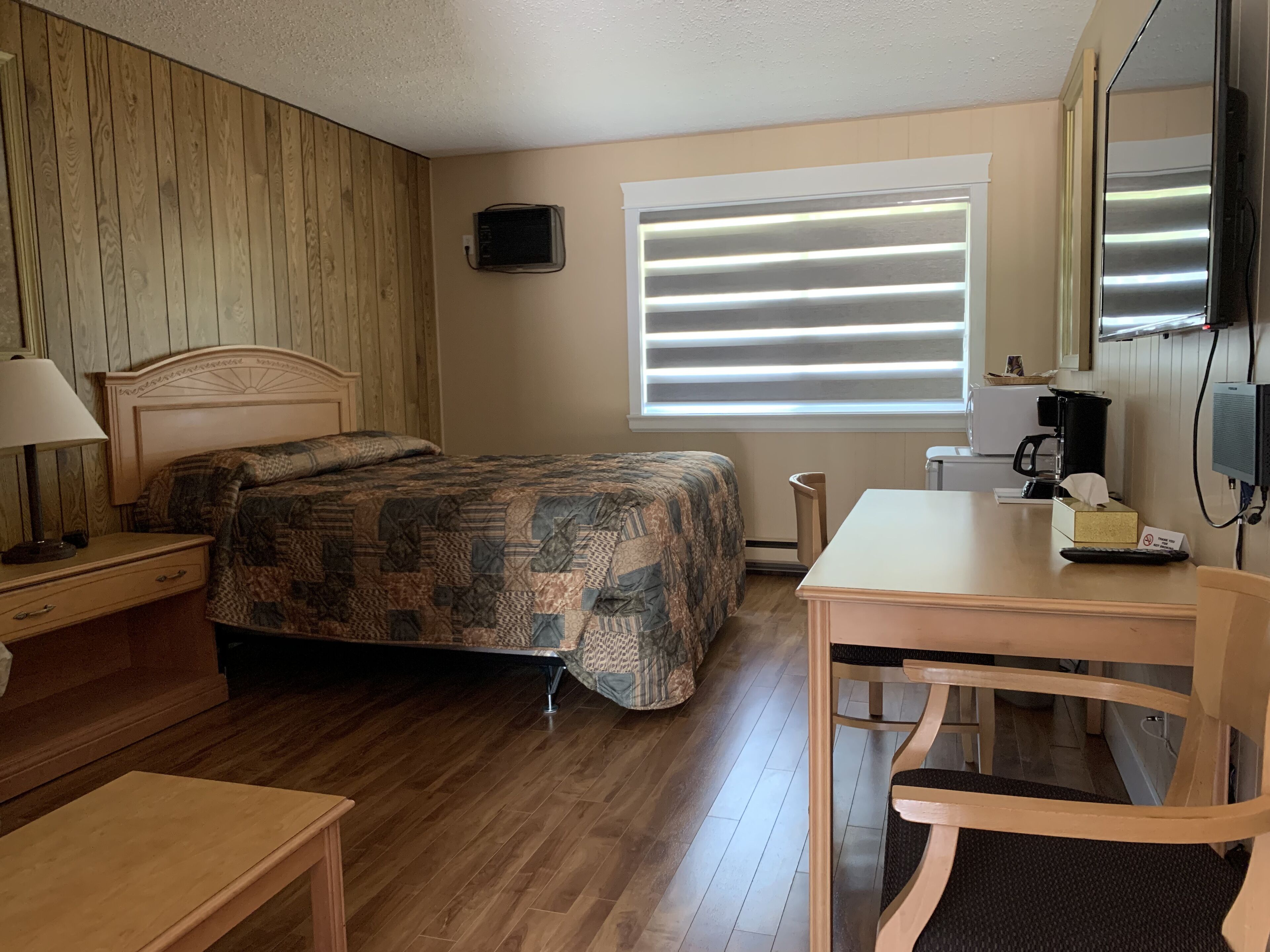 A bedroom with a wooden headboard, a bed with a patterned quilt, a desk with a chair, a window with blinds, and a television on the wall.