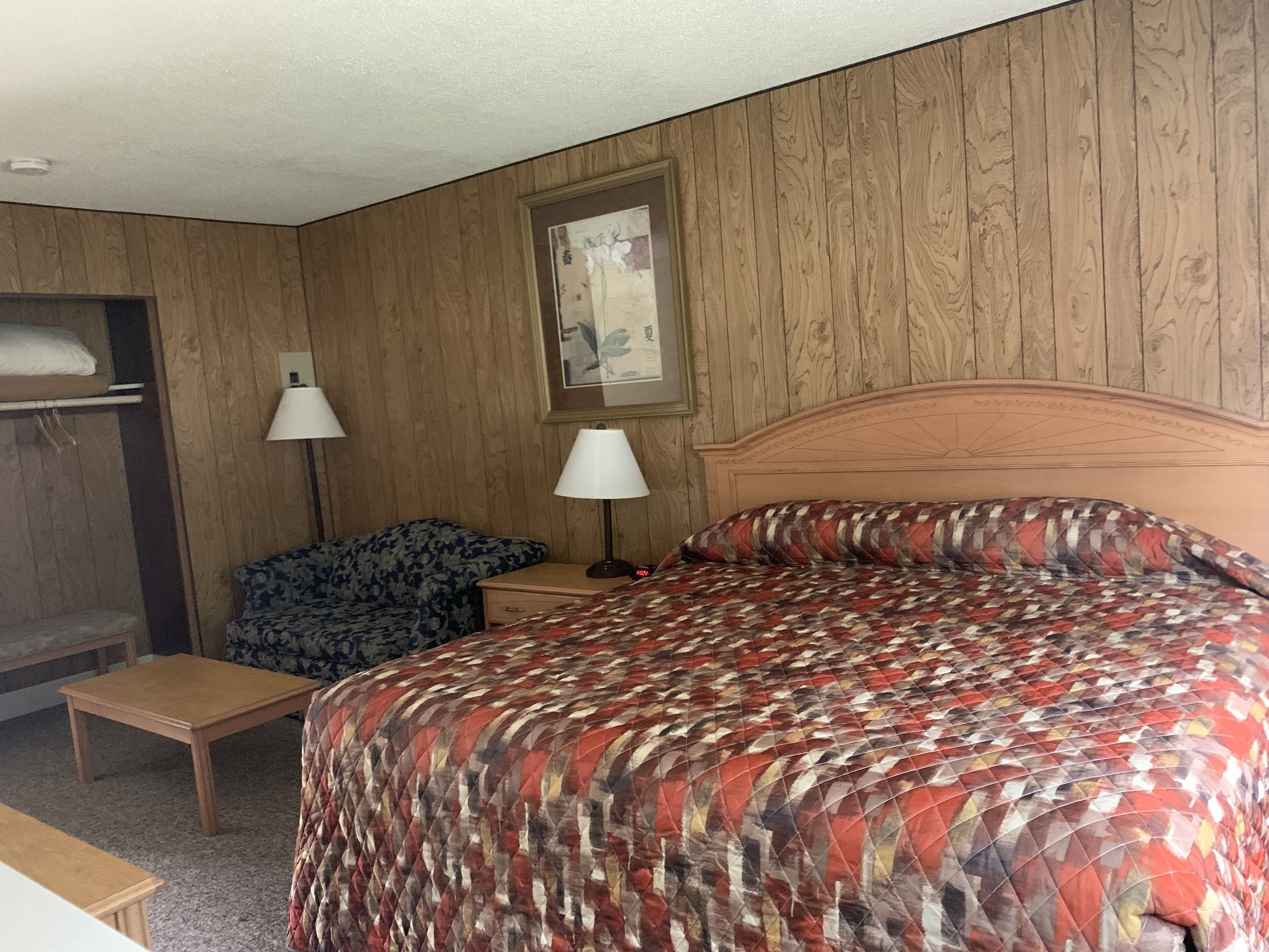 A hotel room with a wooden paneled wall, a bed with a patterned bedspread, a nightstand with a lamp, a small table, and a chair.