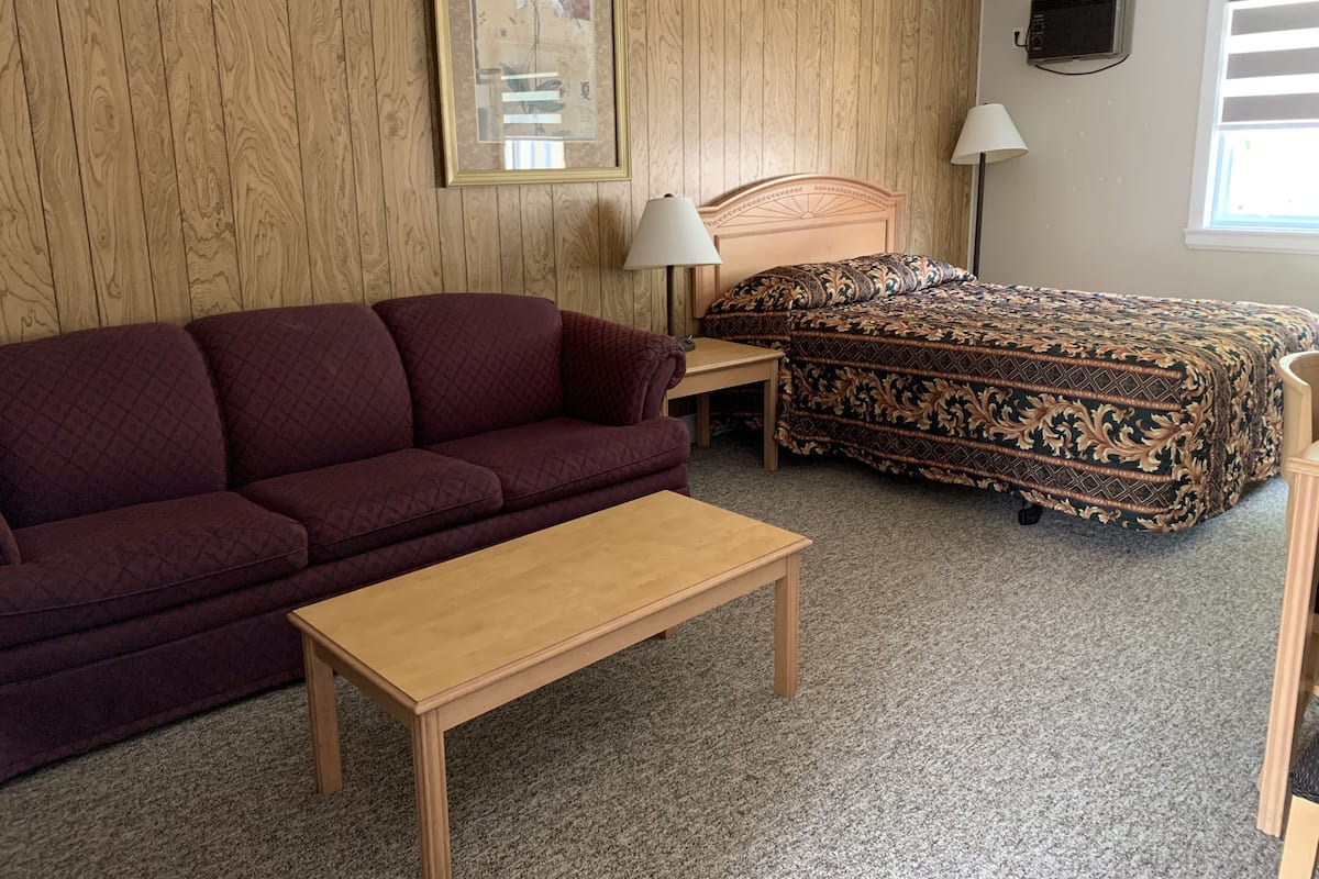 A room with a wooden panel wall, a bed with a patterned bedspread, a red sofa, a wooden table, and two wall-mounted lamps.