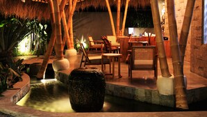 An outdoor dining area with bamboo structures, seating, and a water feature.