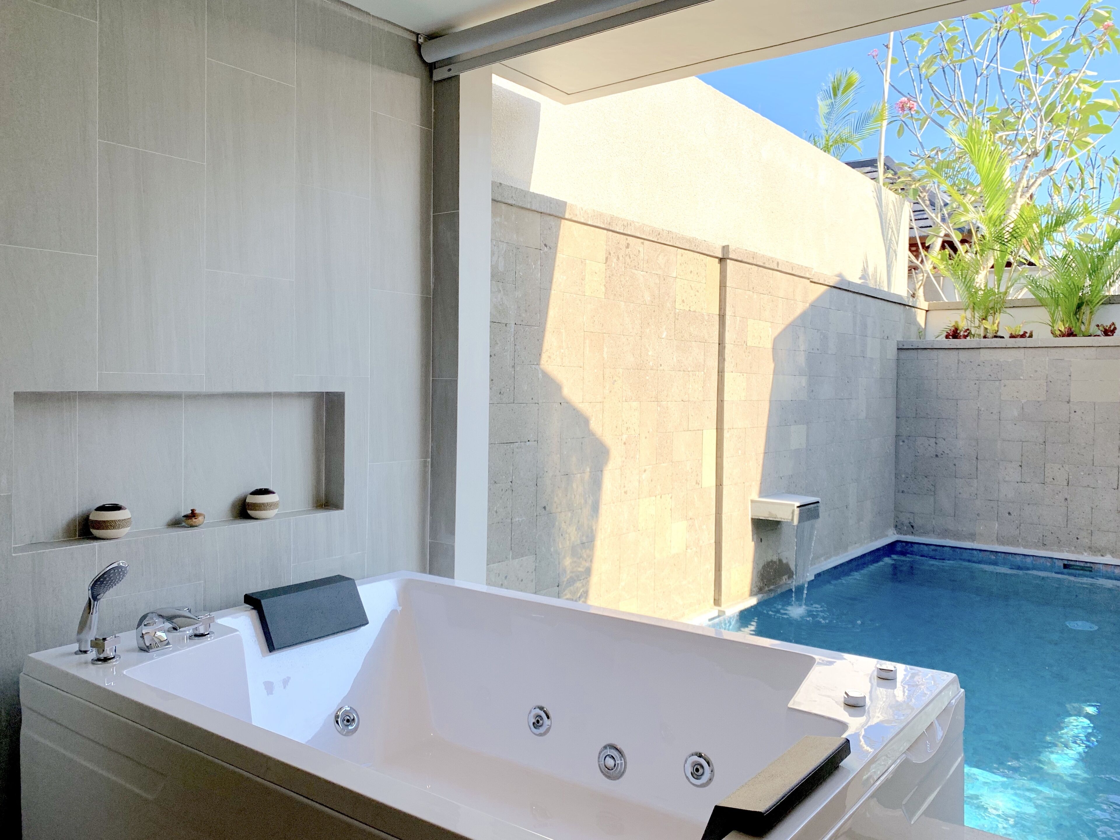 A modern bathroom with a large bathtub, a shower, and a small sink. There are decorative items on the wall and a view of a swimming pool outside.