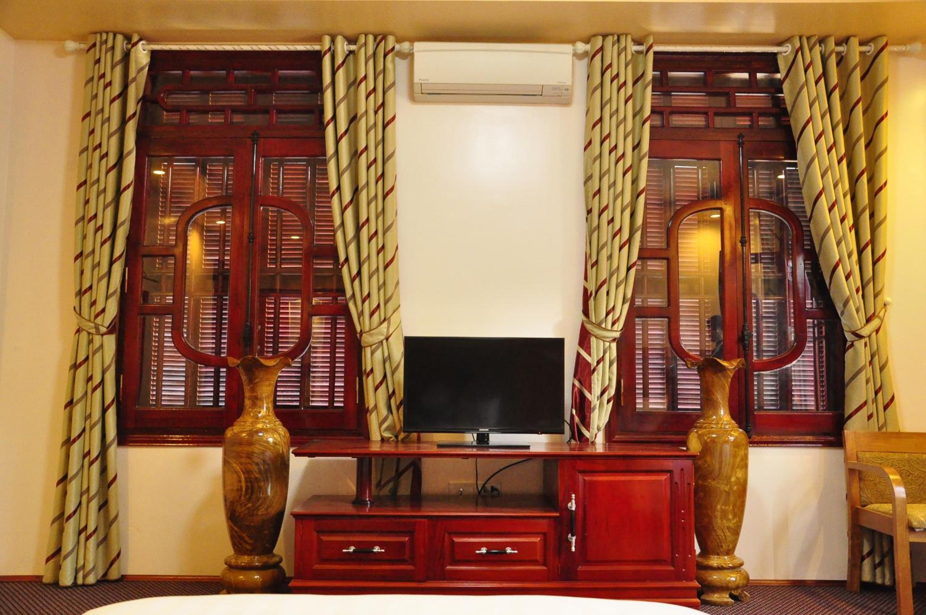 A hotel room with a TV on a wooden cabinet, two large vases, and curtains with a geometric pattern.