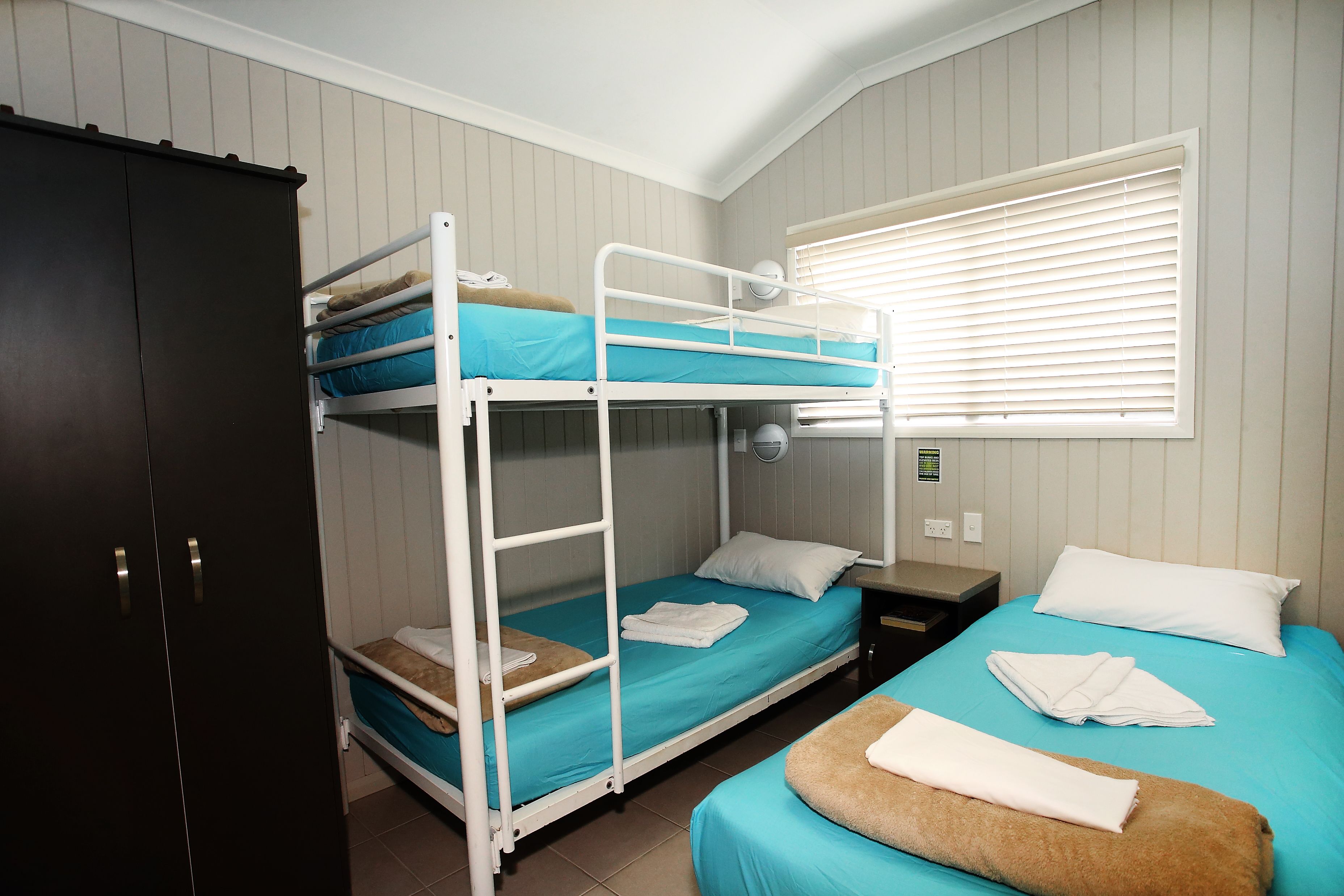 A bunk bed room with a wardrobe, a bedside table, and a window with blinds.