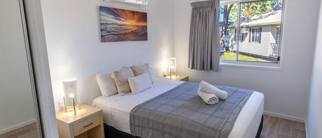 A neatly arranged bedroom with a bed, bedside lamps, a painting, and a window with curtains.