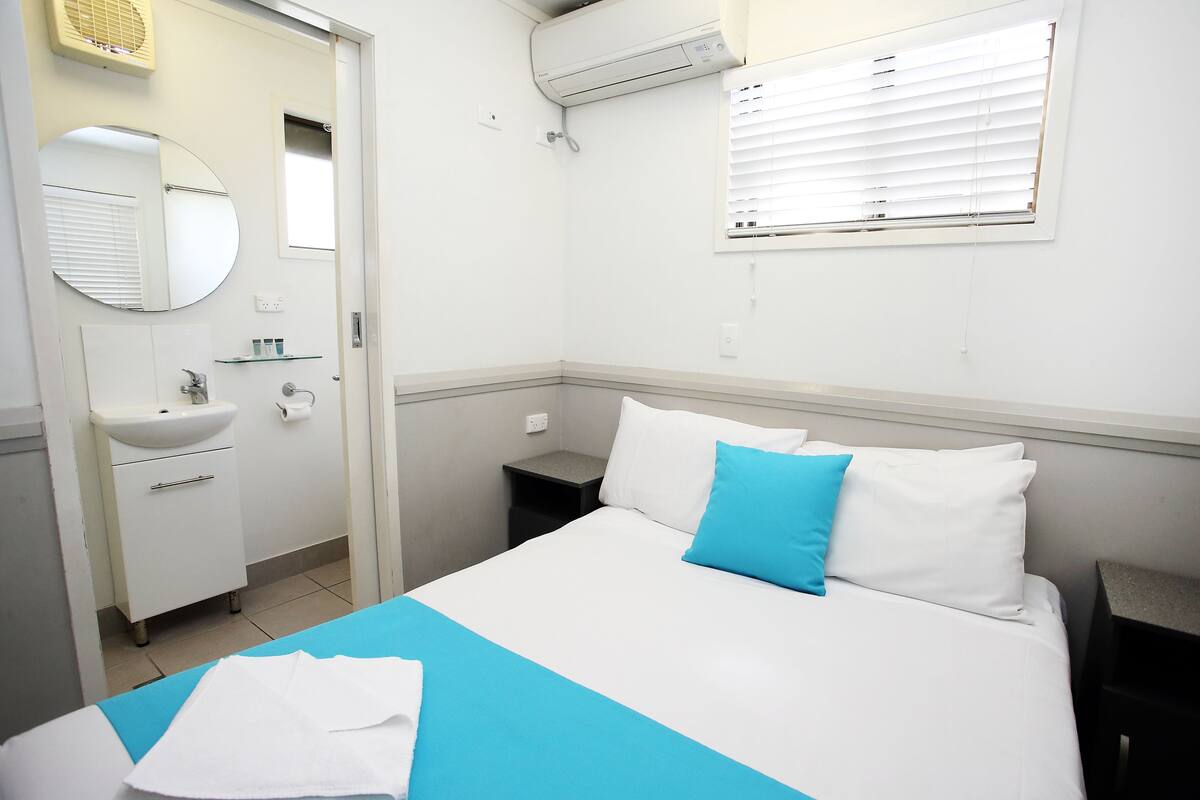 Una habitación de hotel con una cama, una mesita de noche, un pequeño tocador, un lavabo blanco, un espejo y una ventana con persianas.
