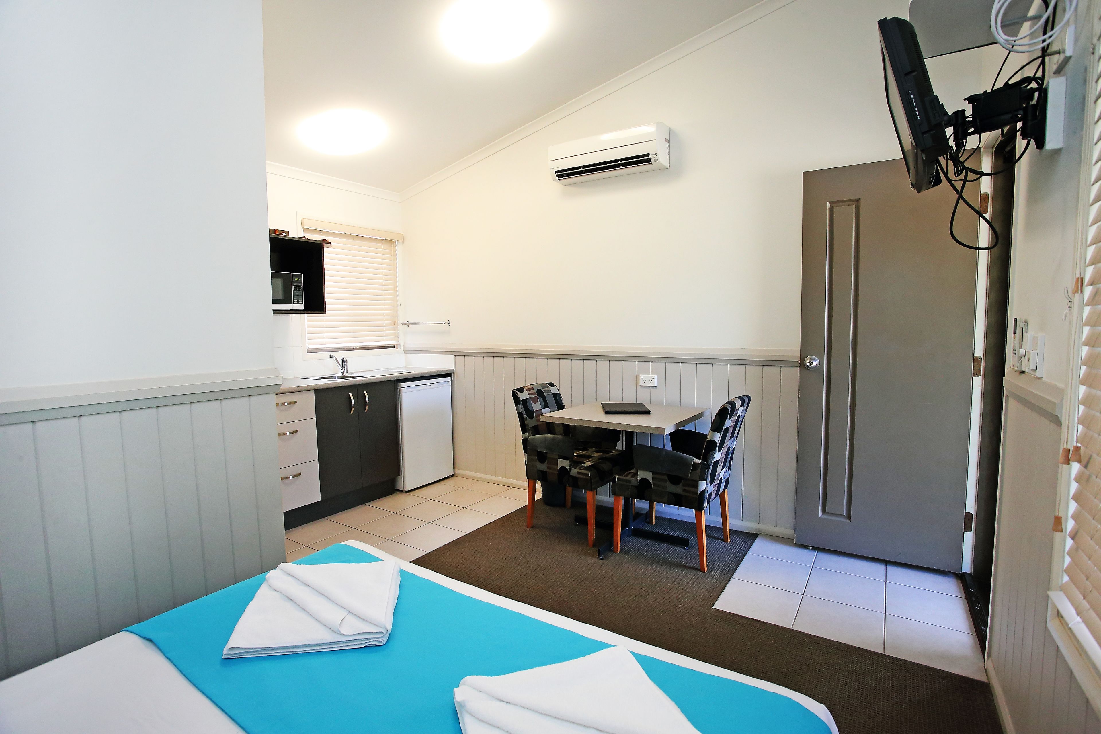 A compact room with a bed, a small kitchenette, a dining table with chairs, and a door.