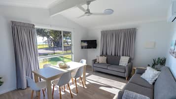 A modern living room with a dining area, grey sofas, a flat-screen TV, and a view of a park.