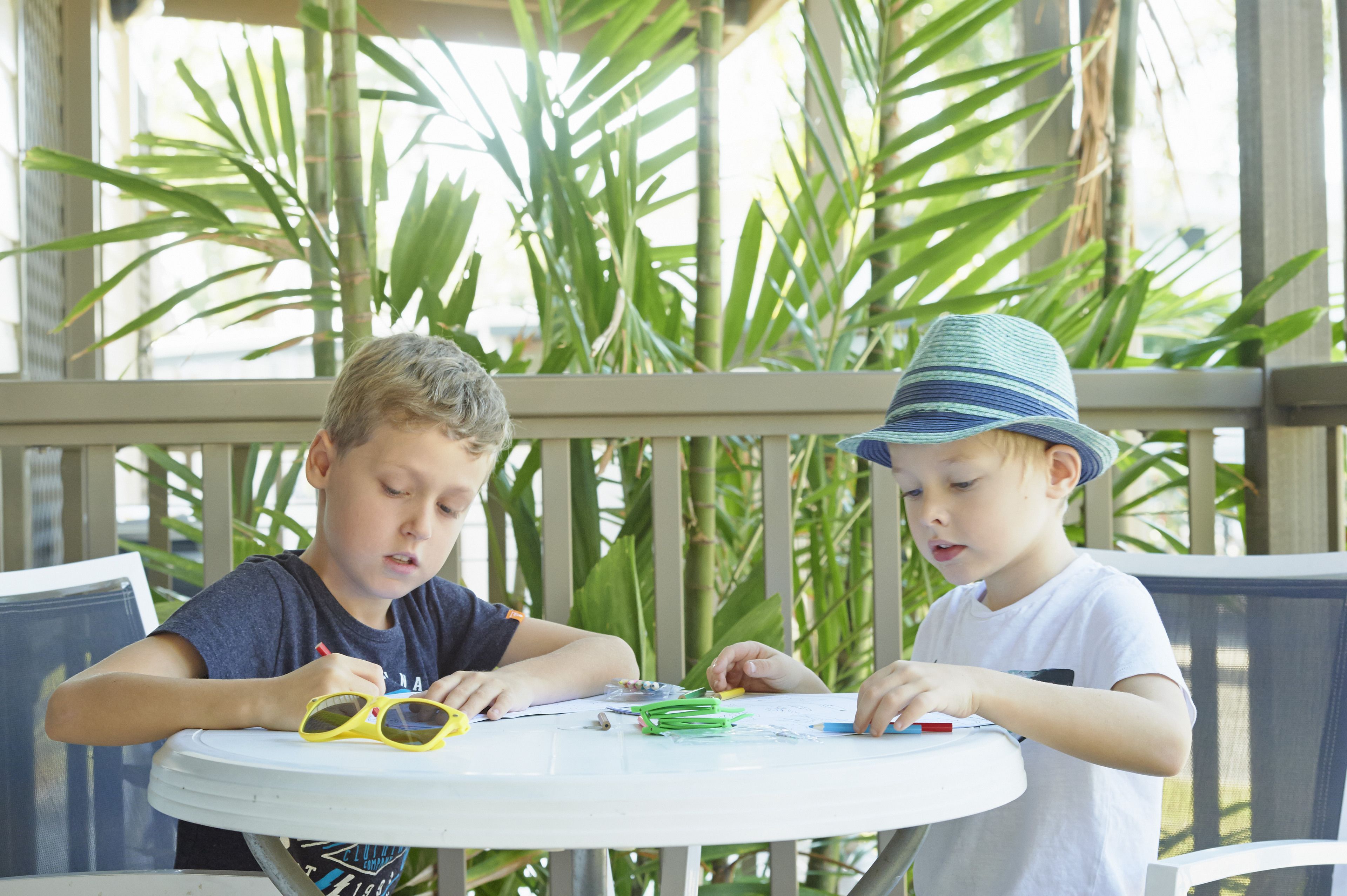 Twee kinderen aan een ronde tafel met een wit tafelkleed, omringd door groene planten.