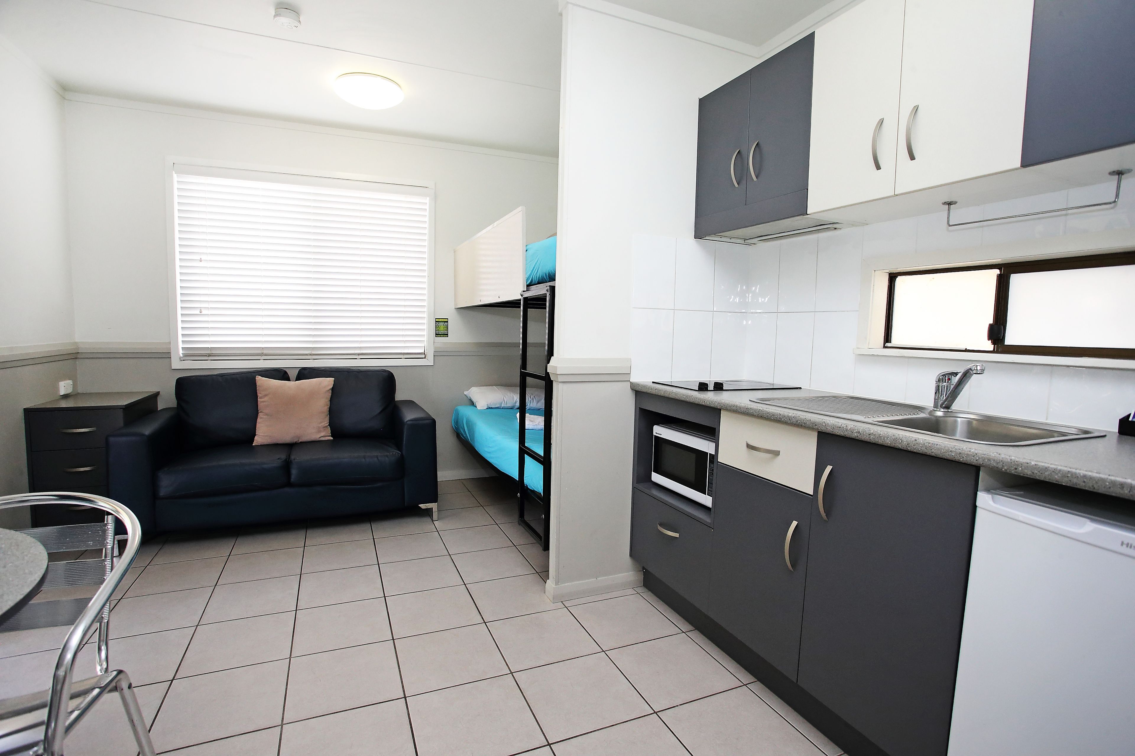 A compact kitchen with a built-in microwave, a sink, and a dishwasher. There is a small dining area with a table and chairs. A bunk bed with a blue mattress is visible in the background.