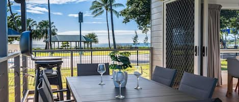 Un espacio al aire libre cubierto para comer, con una mesa y sillas, vista al mar y una palmera de fondo.