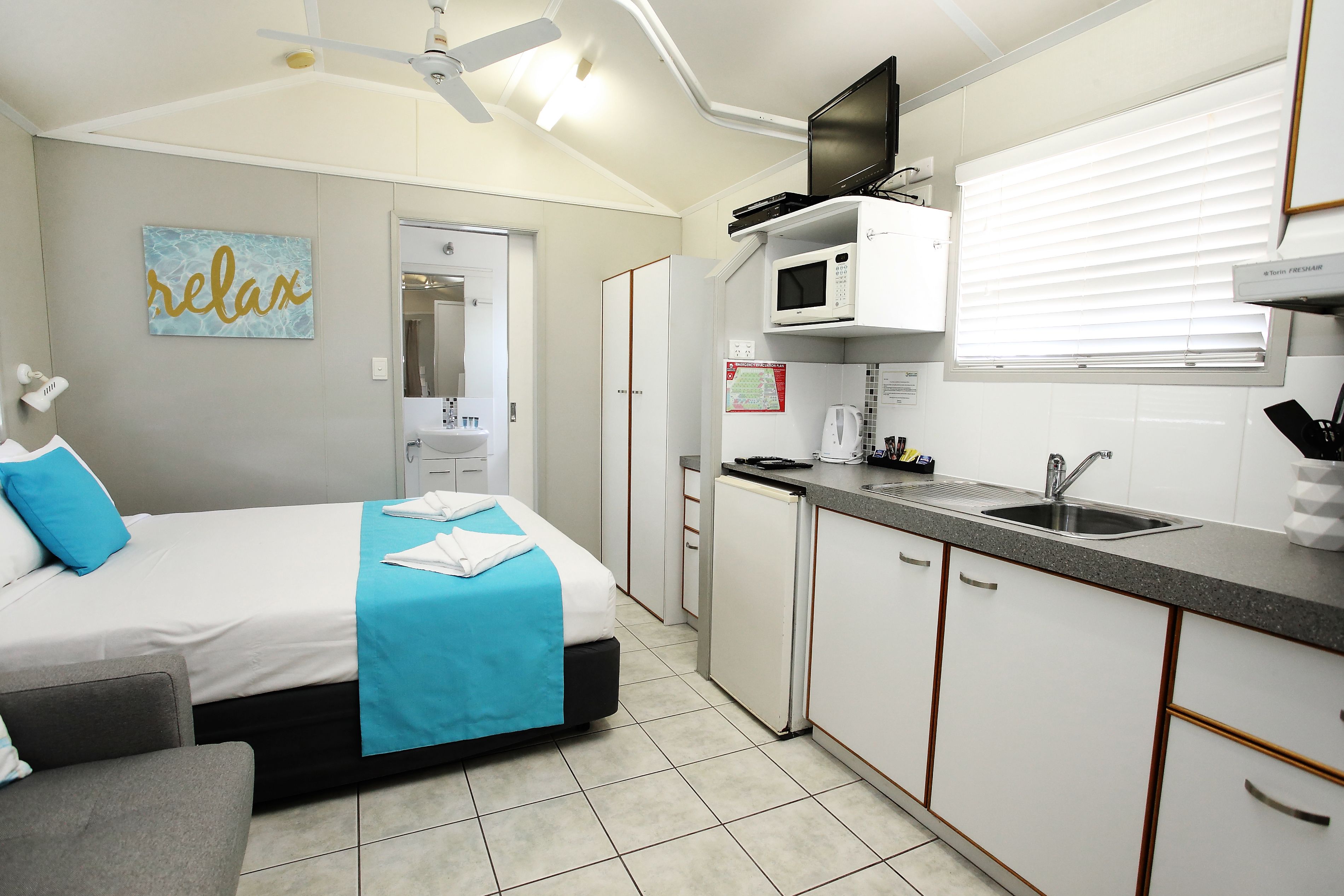 Une chambre d’hôtel de taille réduite, comprenant un lit, une petite kitchenette, un ventilateur de plafond et une salle de bain visible à travers une porte ouverte.