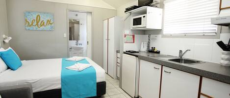 Une chambre d’hôtel de taille réduite, comprenant un lit, une petite kitchenette, un ventilateur de plafond et une salle de bain visible à travers une porte ouverte.