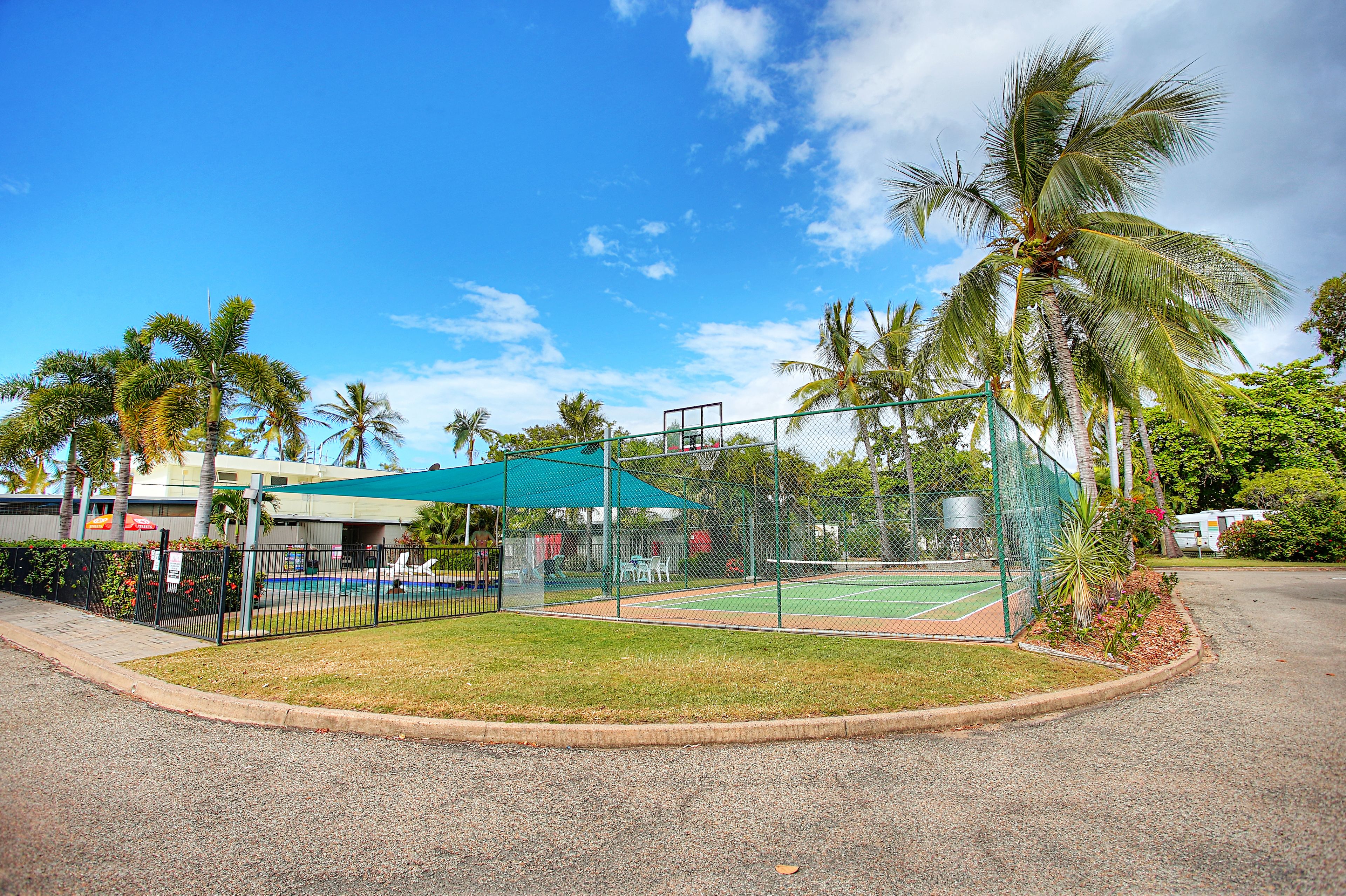 A fenced tennis court with a green canopy, surrounded by palm trees and a building with a green roof.