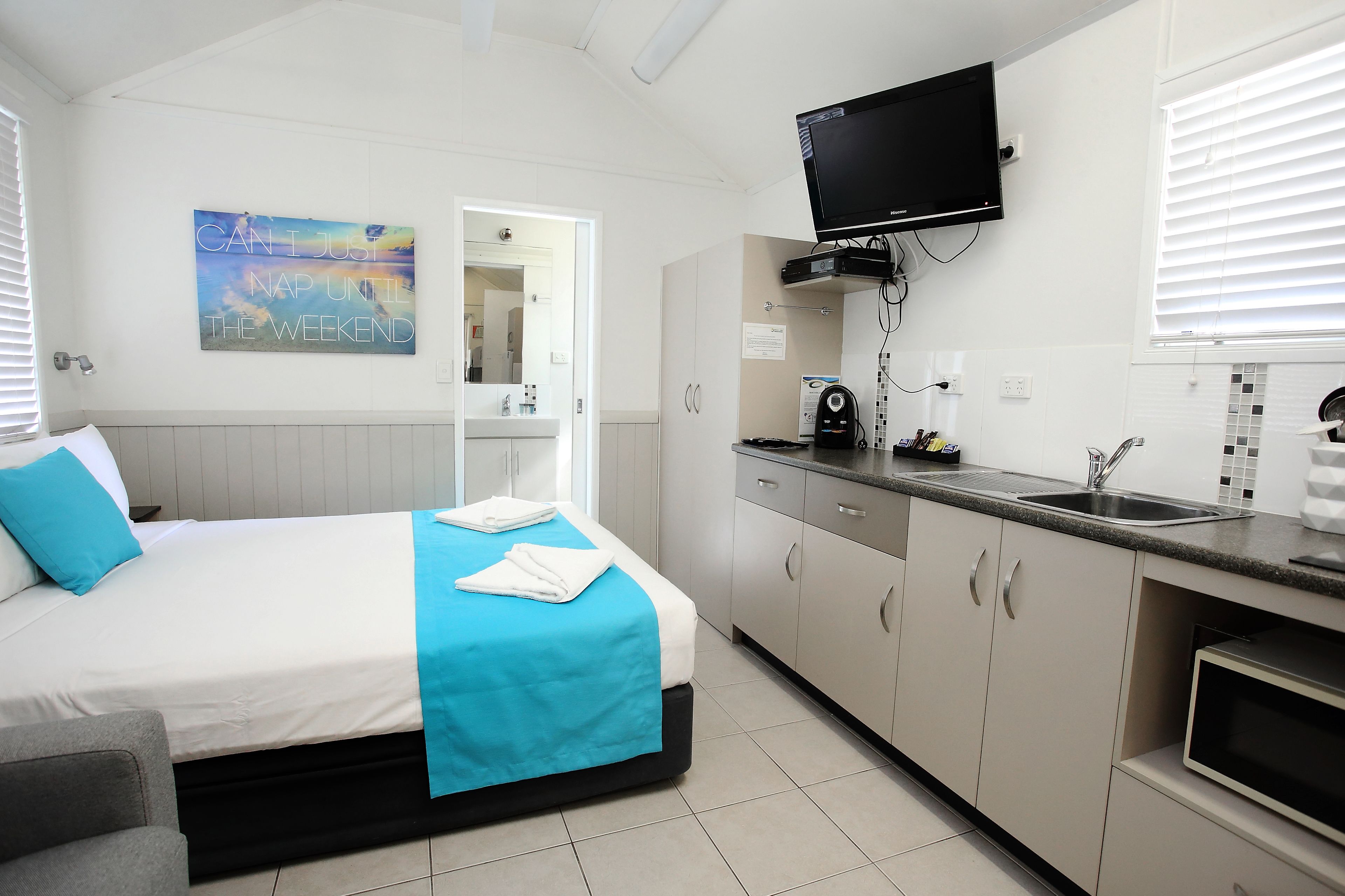 A hotel room with a bed, a TV mounted above the bed, a kitchenette with a sink, and a bathroom visible through an open door.