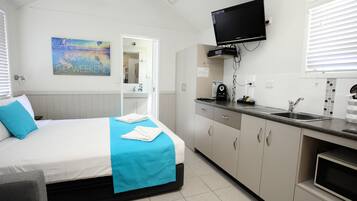 A hotel room with a bed, a TV mounted above the bed, a kitchenette with a sink, and a bathroom visible through an open door.