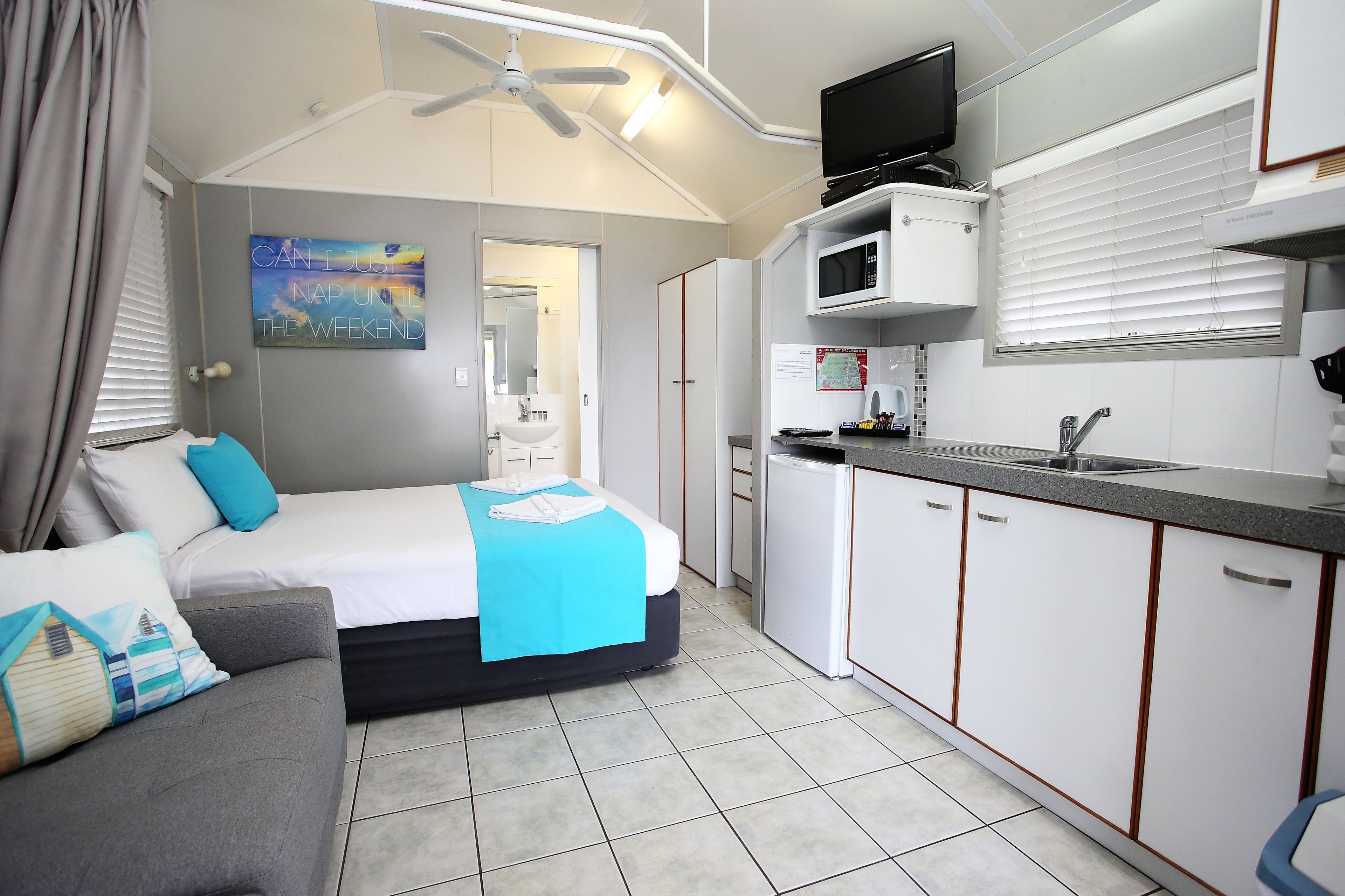 A compact room with a bed, a grey sofa, a kitchenette, and a ceiling fan.