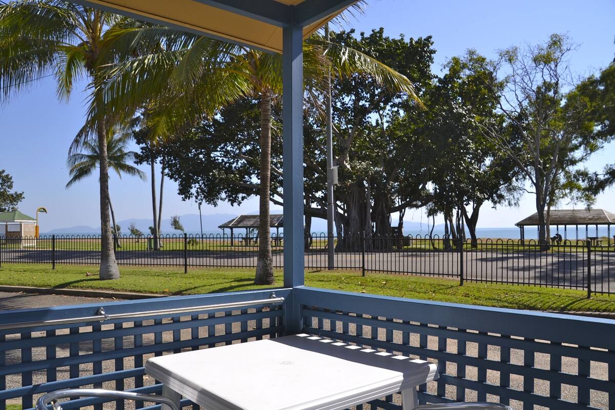 Un área de estar techada con una mesa, rodeada por una valla y palmeras, con vista a una playa y un parque.