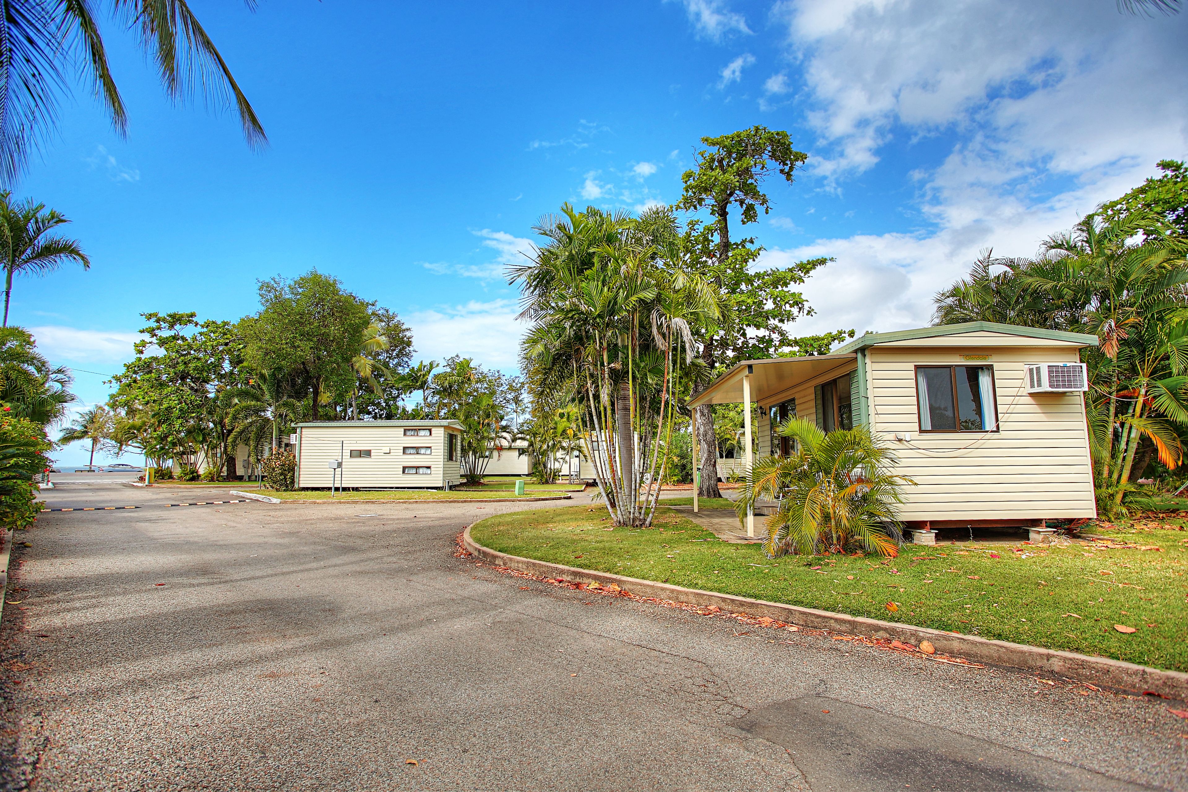 Un barrio residencial con una calle pavimentada, una casa pequeña con techo verde y palmeras alrededor.
