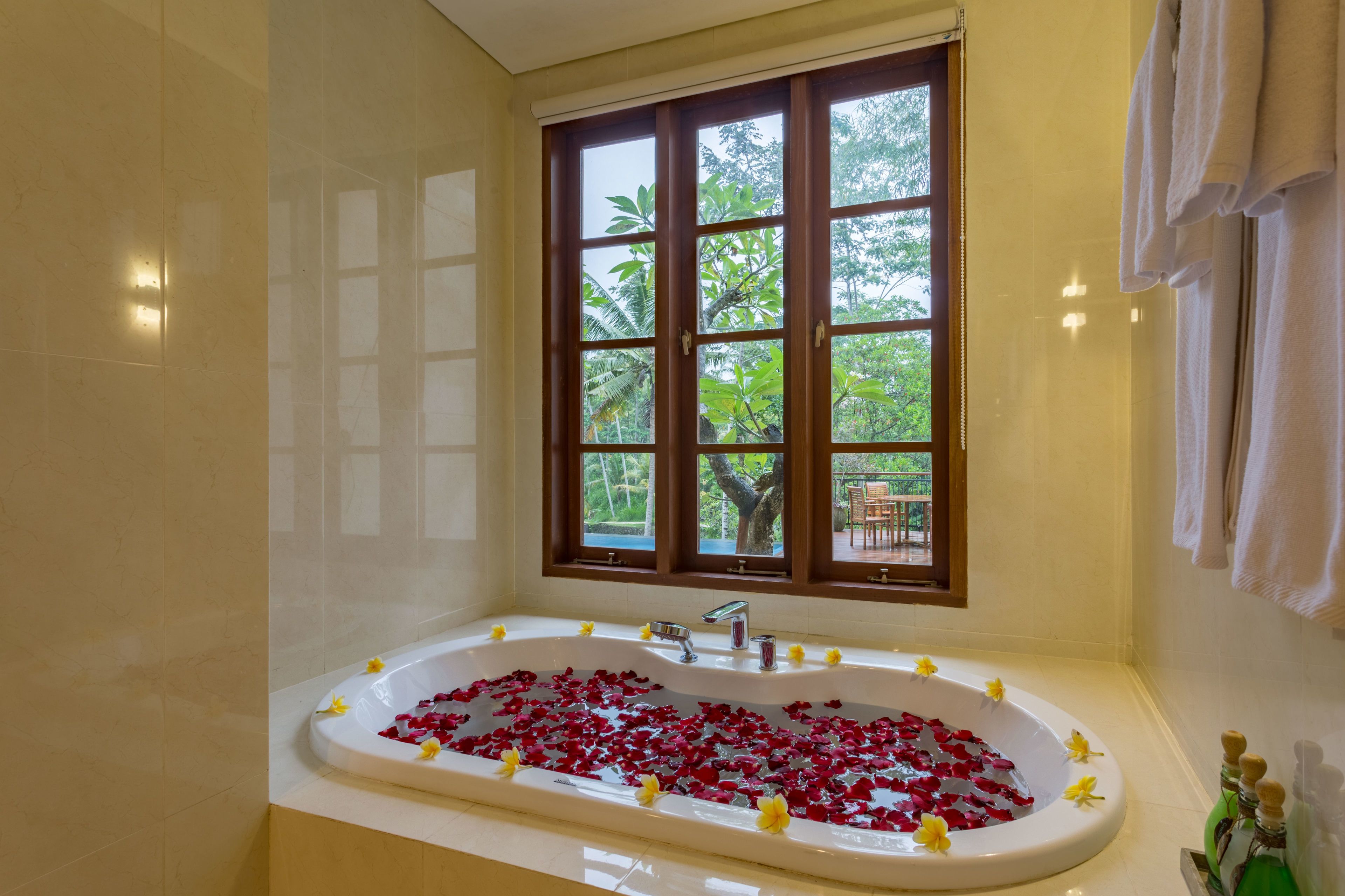 A bathroom with a bathtub filled with rose petals, a window with a view of greenery, and a towel rack.