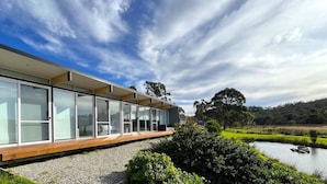 Un edificio moderno con amplios ventanales de vidrio y una terraza de madera, ubicado junto a una masa de agua y rodeado de vegetación.