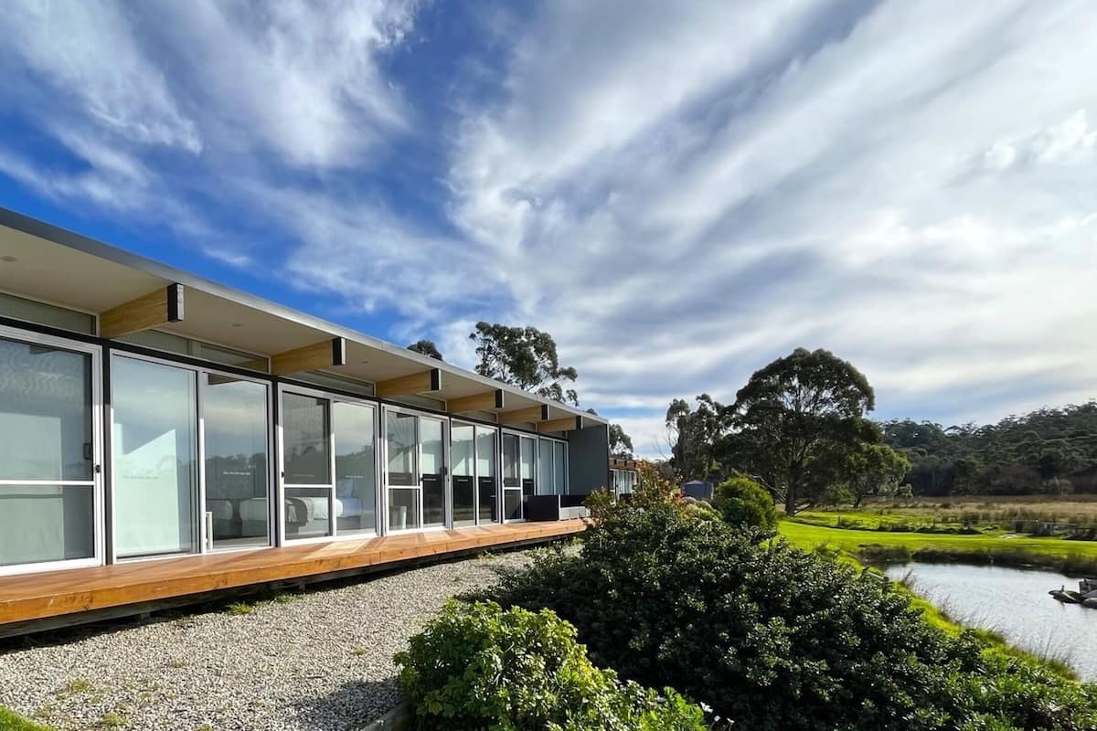Un edificio moderno con amplios ventanales de vidrio y una terraza de madera, ubicado junto a una masa de agua y rodeado de vegetación.