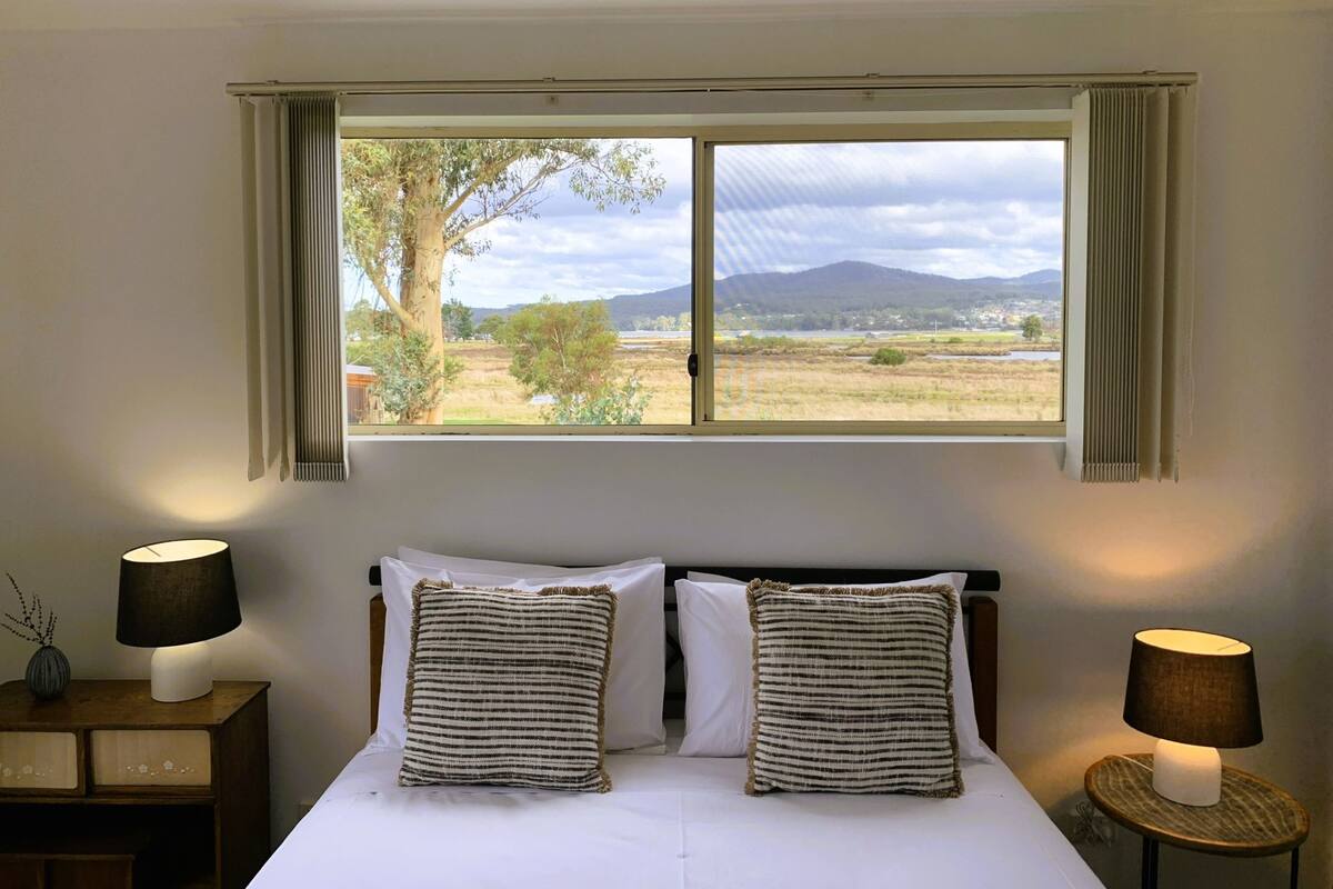 Un dormitorio con cama, dos lámparas de noche, una ventana con cortinas y vista a un paisaje.