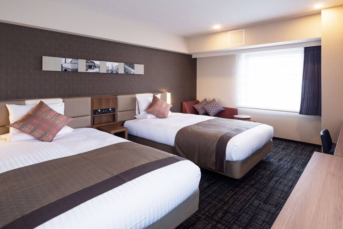 Ein Hotelzimmer mit zwei Betten, einem Schreibtisch, einem Stuhl, einem Fernseher und einem Fenster mit Vorhängen.