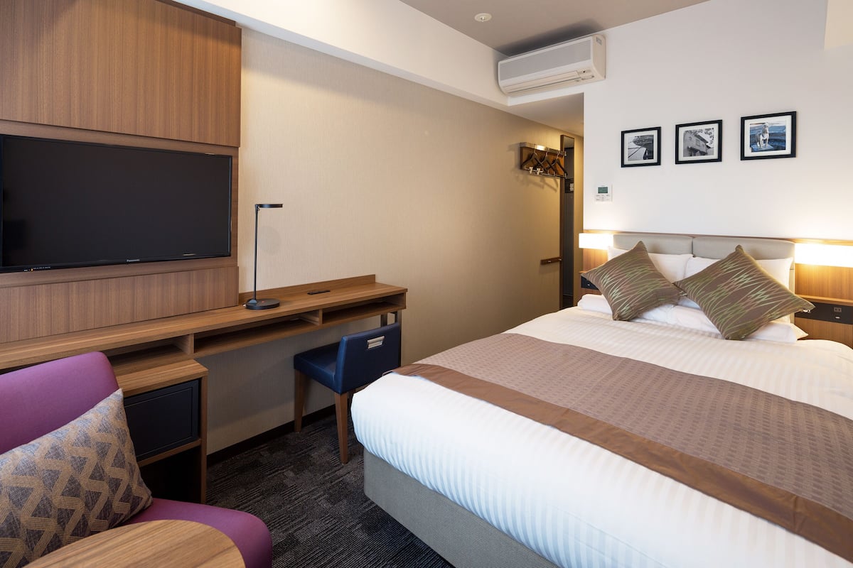 Ein Hotelzimmer mit Bett, Schreibtisch, Stuhl, Flachbildfernseher und Wandklimaanlage.