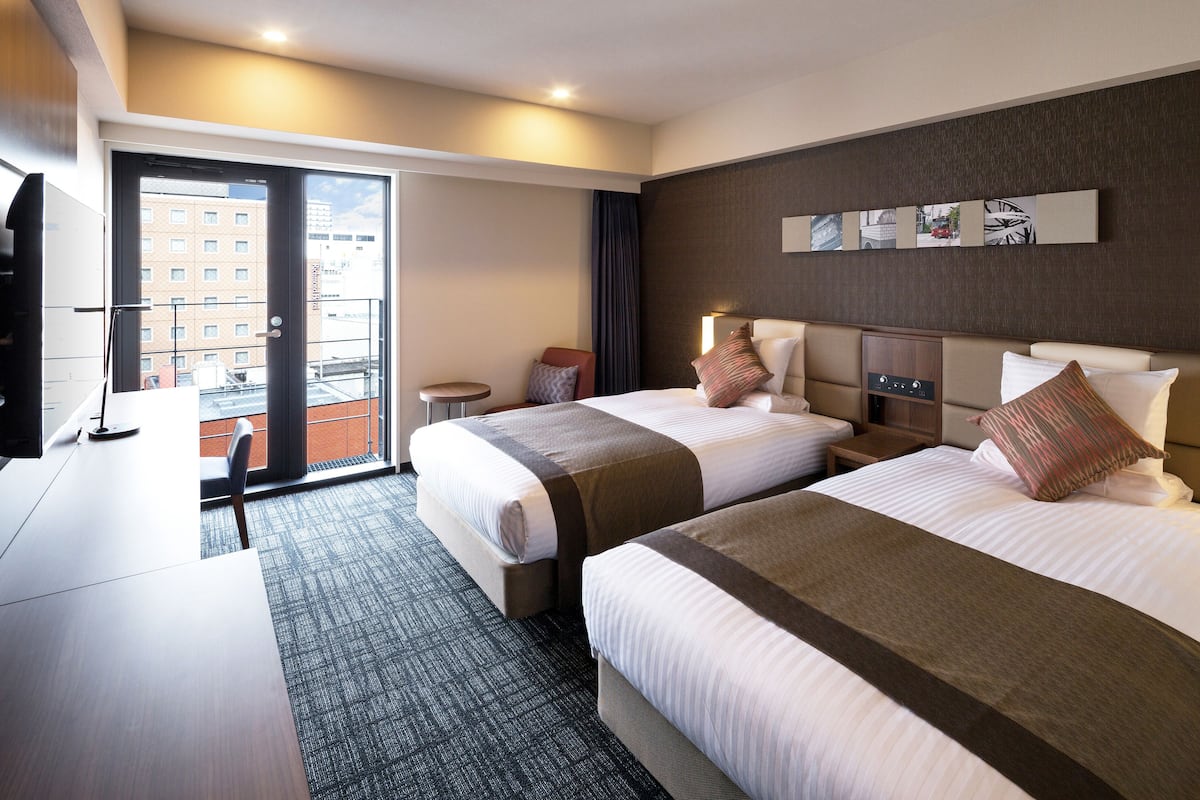 Ein Hotelzimmer mit zwei Betten, einem Schreibtisch und Blick auf die Stadt durch ein großes Fenster.