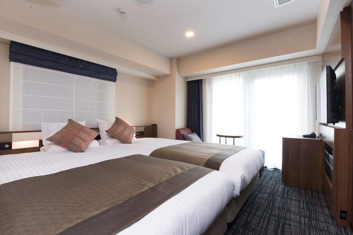 Ein Hotelzimmer mit zwei Betten, einem Fernseher, einem kleinen Tisch und einem Fenster mit Vorhängen.