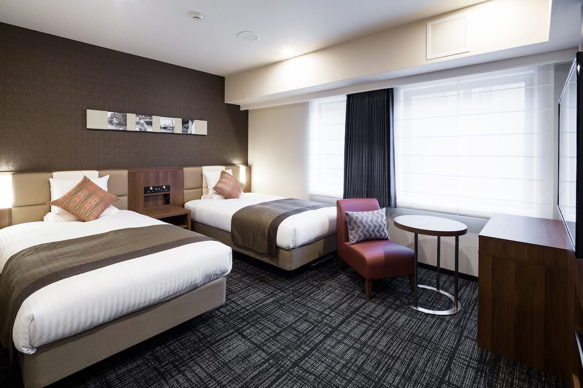 Ein Hotelzimmer mit zwei Betten, einem kleinen runden Tisch, einem roten Sessel und einem großen Fenster mit Vorhängen.