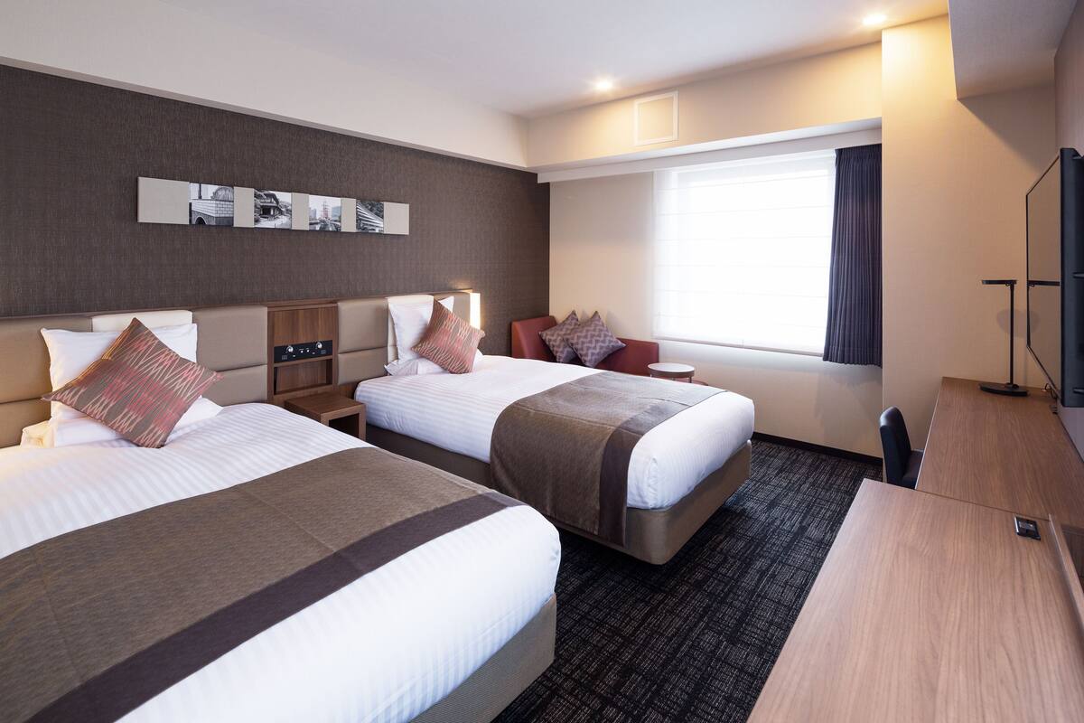 Ein Hotelzimmer mit zwei Betten, einem Schreibtisch, einem Stuhl, einem Fernseher und einem Fenster mit Vorhängen.
